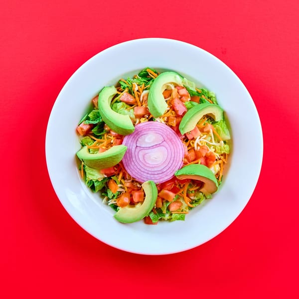 A vibrant Mexican salad with lettuce, tomatoes, avocado, shredded cheese, and red onion on a white plate.
