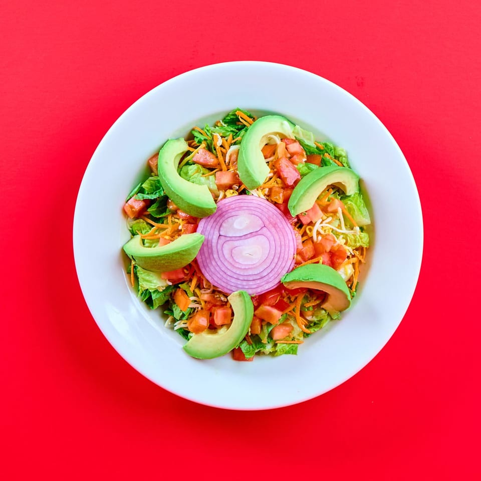 A vibrant Mexican salad with lettuce, tomatoes, avocado, shredded cheese, and red onion on a white plate.